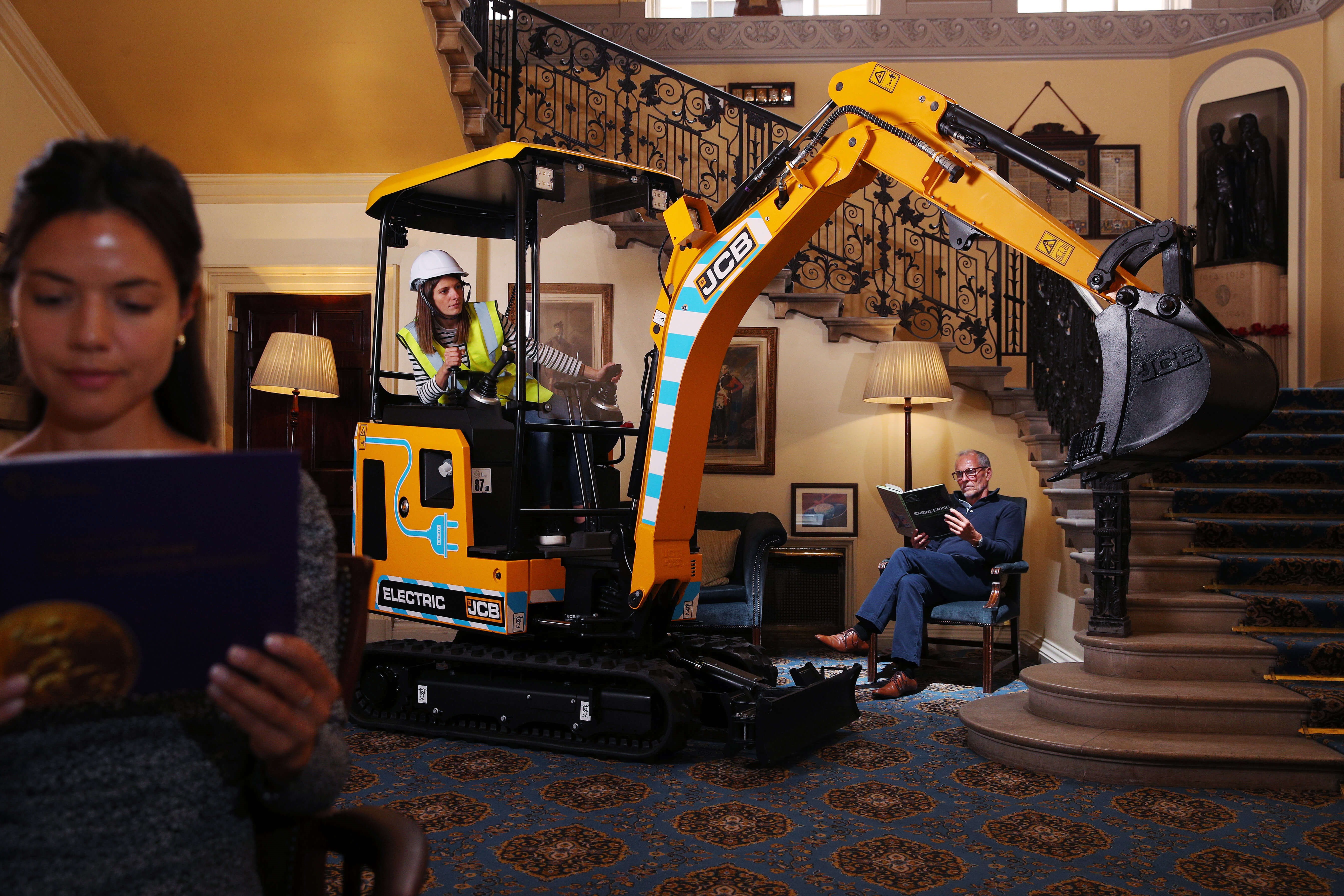 JCB electric digger being operated indoors, next to two people reading and unaware of its presence