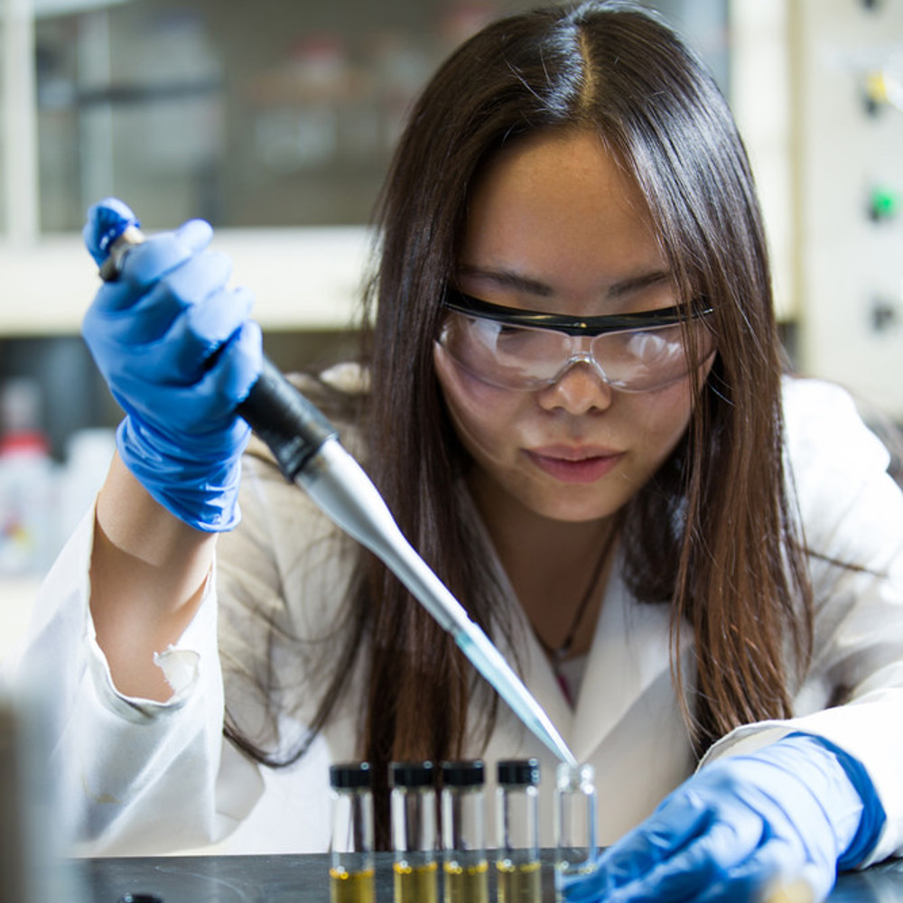 Engineer wearing a lab coat is using a pipette to drop a substance into a small container.
