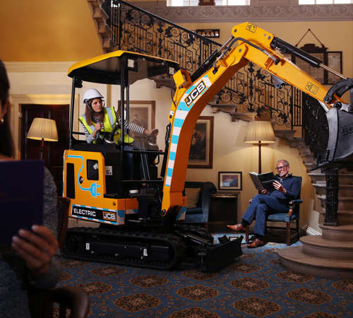 JCB electric digger being operated indoors, next to two people reading and unaware of its presence