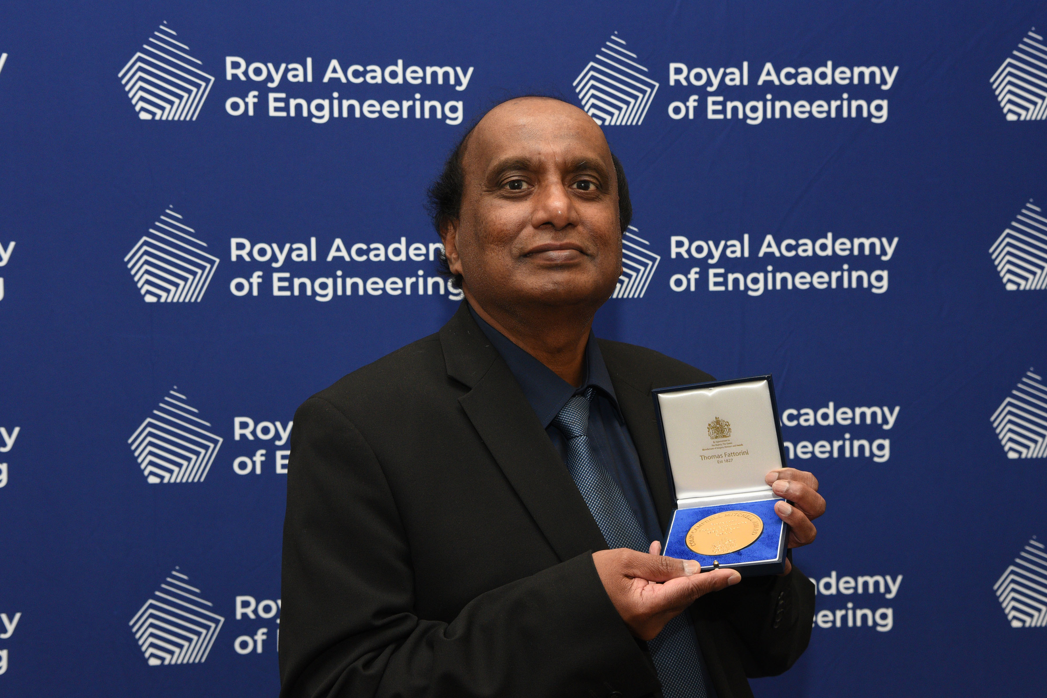 Professor Mohan Edirisinghe OBE FREng stood holding the Colin Campbell Mitchell Award in front of a blue screen with the Royal Academy of Engineering logo