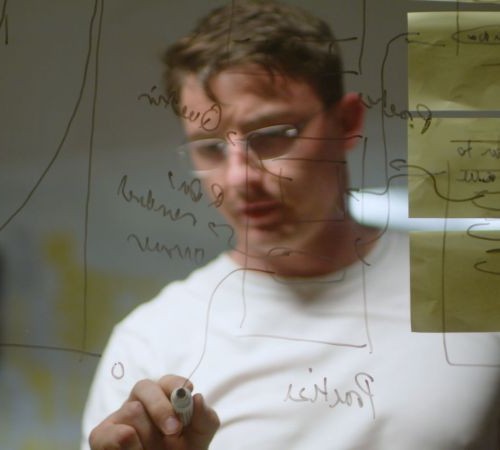 A man draws mathematical graphs and equations on a clear plastic screen in front of the camera.