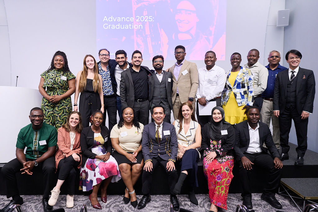 Advance 2025 awardees stand on a stage in a large huddle, smiling towards the camera.