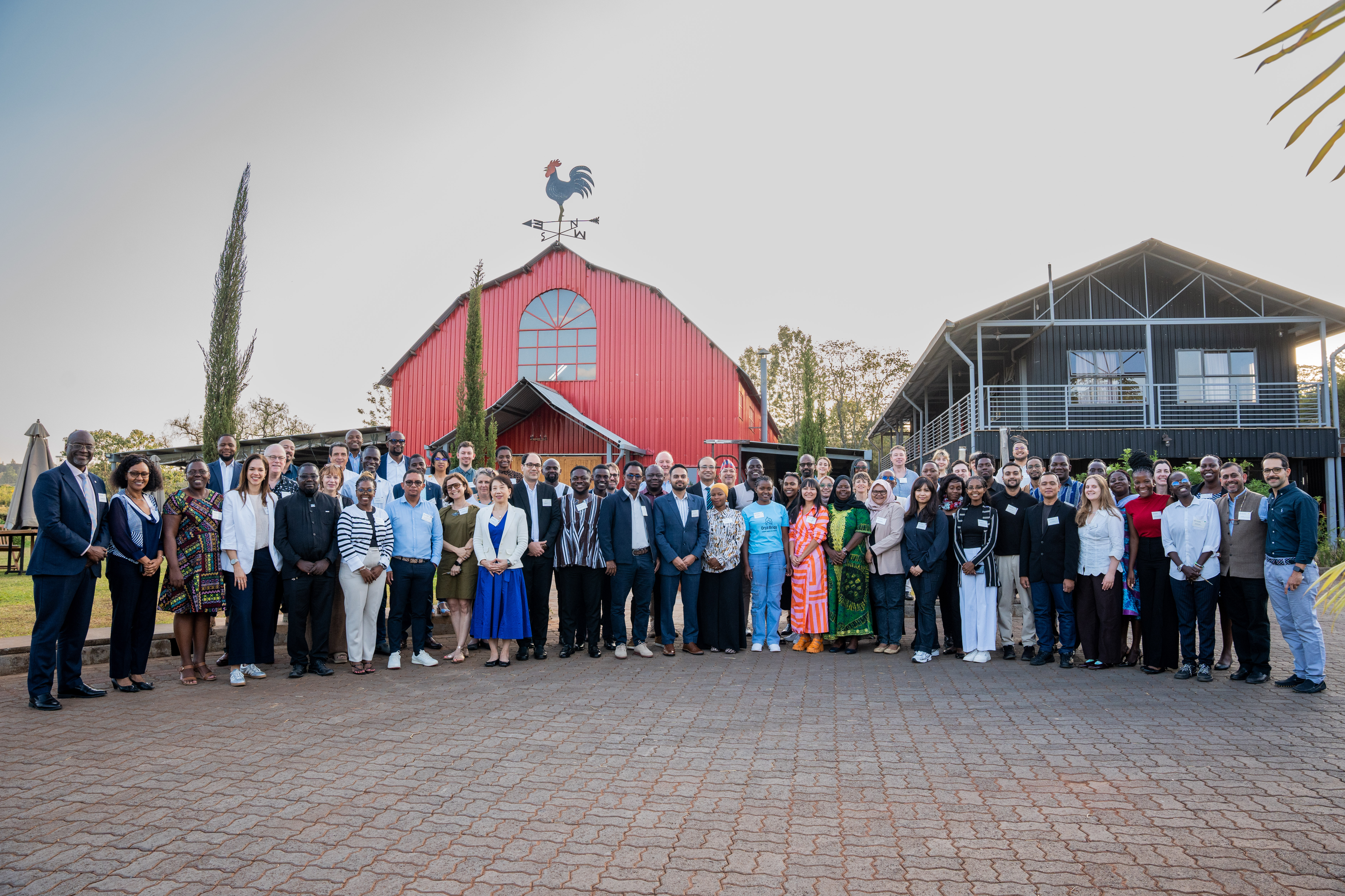 A diverse group of roughly 65 people stand outside a big red barn, smiling at the camera