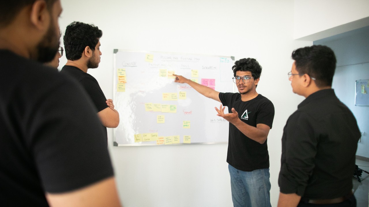 Four men discuss with one another, while one of them points to a whiteboard.
