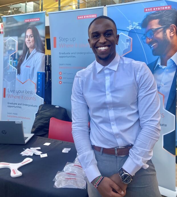 Engineer smiling, standing in front of a BAE Systems display.